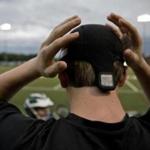 A Westwood Youth Lacrosse player adjusts a Checklight sensor, with its display panel visible above his neck,  before  putting on  his helmet. 
