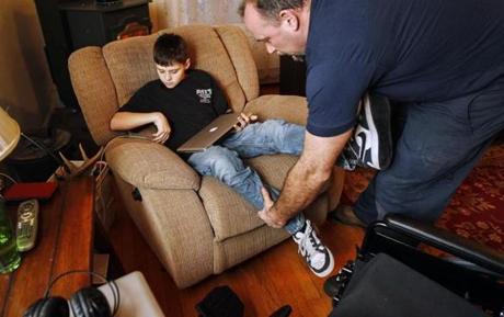 Dan Dugan, 13, hooked up his laptop as his dad, Pat Dugan, adjusted his legs at their home in Seabrook, N.H. Dan has weakness in his legs and is making slow improvements but needs a wheelchair.
