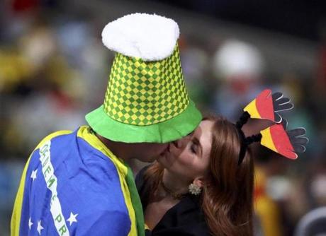 A man with a Brazilian flag draped over his shoulders kisses a woman dressed in the colors of the German flag. 
 
