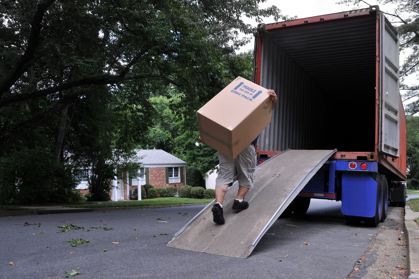 An unidentifiable person is carrying a heavy moving box up a ramp into the back of a moving truck (altered features, not recognizable), on a summer day in typical suburban America.