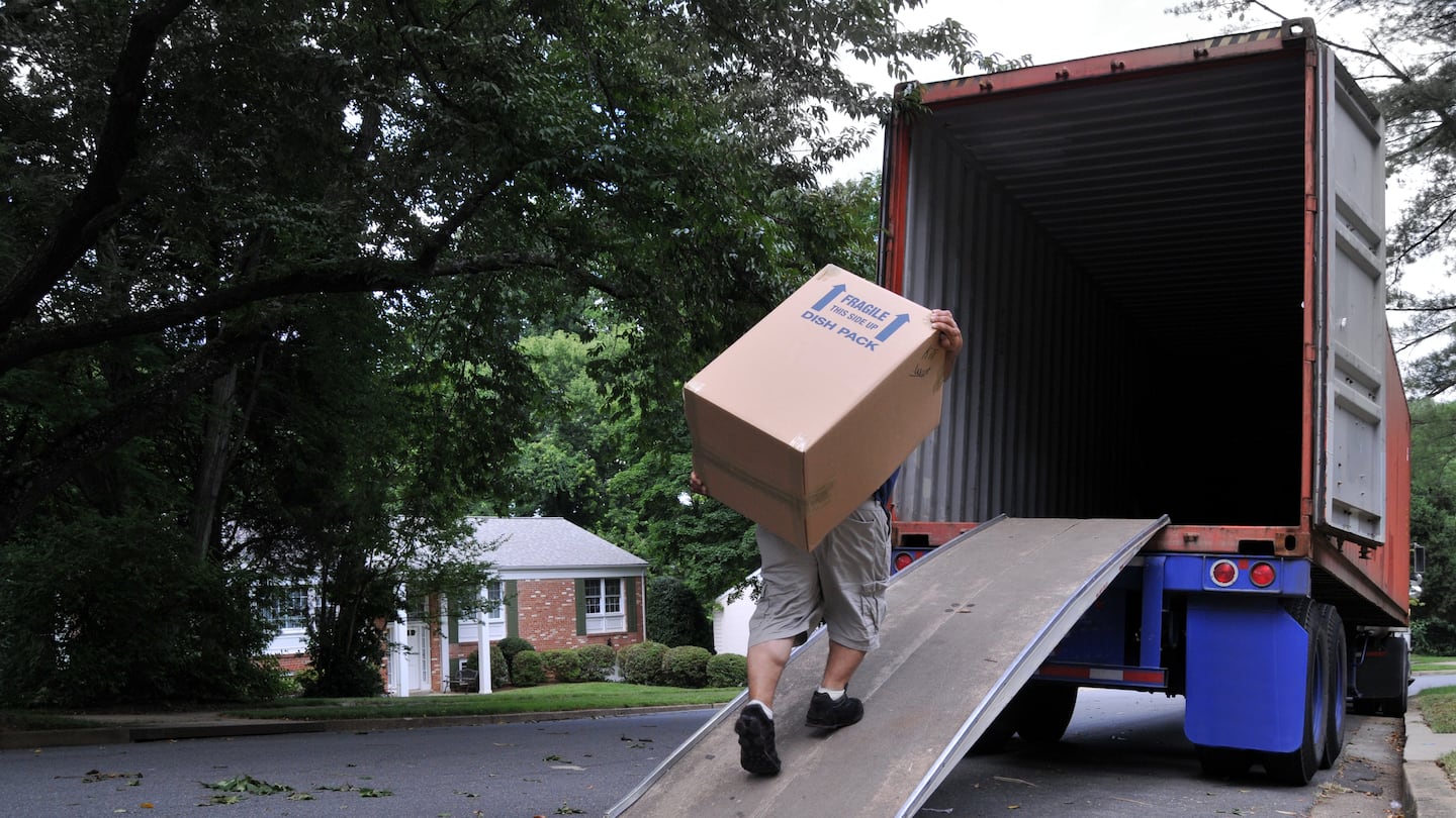 An unidentifiable person is carrying a heavy moving box up a ramp into the back of a moving truck (altered features, not recognizable), on a summer day in typical suburban America.