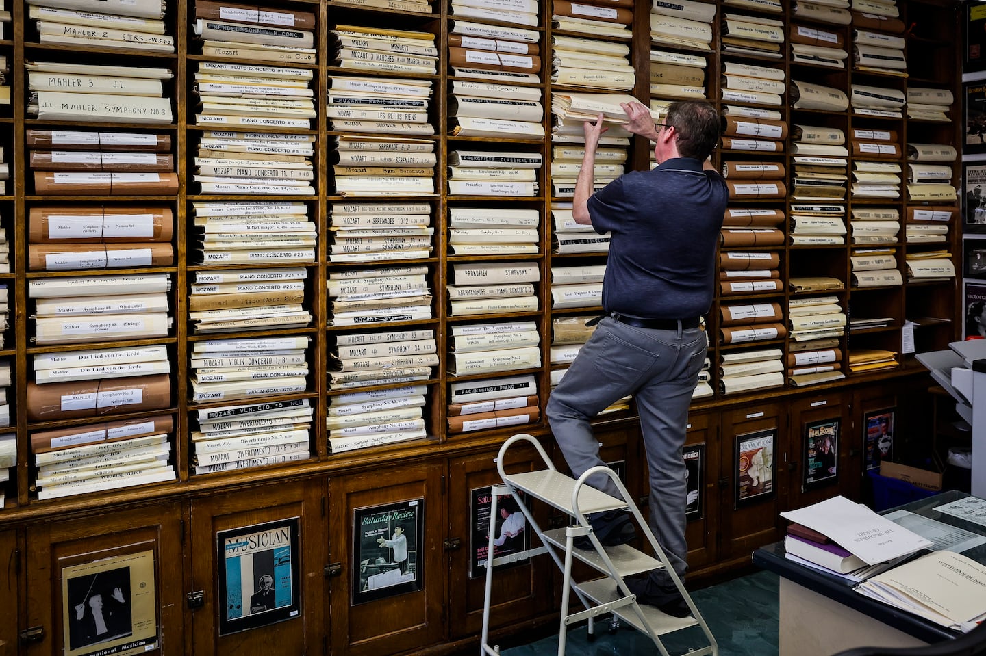 Principal librarian Wilson Ochoa reaches for scores stored in floor-to-ceiling wooden shelving at the Boston Symphony Library, where hundreds of orchestral works by composers like Mahler and Mozart are archived in white folios. Orchestral librarians are responsible for managing and preparing all musical scores and parts for performances, including marking detailed instructions in individual parts, maintaining the music library, and ensuring proper distribution of materials to conductors and musicians.