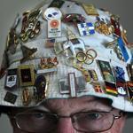 Bob Boehm of Lowell dons his hat adorned with Olympic pins that he wears to Olympic Games and collector shows. Boehm has accumulated some 5,000 pins and other Olympic memorabilia over the past 40 years.  The map on the wall marks the 13 Olympic Games that he has attended. He did not go to Sochi as he had planned due to personal reasons.