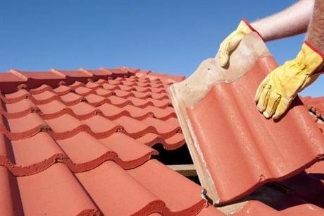 Roof repairs, worker with yellow gloves replacing red tiles or shingles on house with blue sky as background and copy space.
