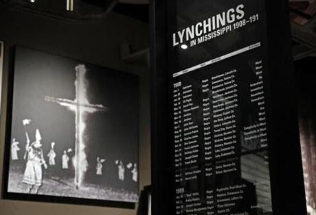 In this Nov. 10, 2017 image, a monolith listing the names, dates and rationale for the lynching of African-American residents rests in the foreground of a photograph of a burning Ku Klux Klan cross on display in the Mississippi Civil Rights Museum in Jackson, Miss. The monolith is one of several that line this gallery with the documented lynchings. Room has been left for updating as needed. This facility is adjacent to the newly built Museum of Mississippi History, that documents the state's rich history and the diversity of its people. Work crews and archivists are putting the final touches on the two museums set to open Dec. 9. (AP Photo/Rogelio V. Solis)
